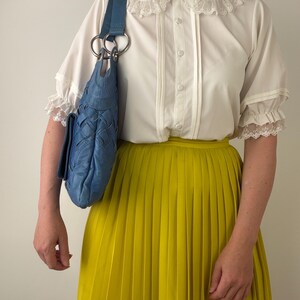 May include: A white short-sleeved blouse with a lace-trimmed Peter Pan collar and button-down front, paired with a bright yellow pleated skirt and a blue woven leather shoulder bag featuring large silver-toned metal rings on the strap.