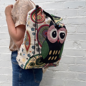 May include: A drawstring backpack with a colourful owl design. The owl has large pink eyes and green wings. The bag features a paisley pattern in red, green, and blue on a cream background. The bag is carried over the shoulder.
