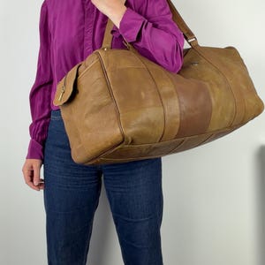 May include: A brown leather duffel bag with a patchwork design. The bag has a zippered side pocket and a shoulder strap. The bag is held by a person wearing a purple shirt and blue jeans.