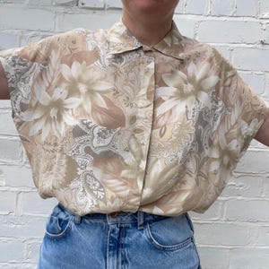 May include: A short-sleeved button-down shirt with a floral pattern in beige, brown, and gray tones. The shirt is paired with blue denim jeans. The floral design includes large flower and paisley motifs.