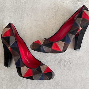 May include: A pair of high-heeled shoes with a geometric pattern in shades of red, blue, brown, and grey. The shoes are made of suede and have a pointed toe.