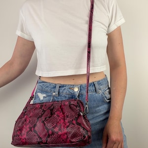 May include: A crossbody bag featuring a vibrant hot pink and black snakeskin pattern. The bag has a structured frame opening, a gathered top design, and a matching thin, adjustable snakeskin-patterned shoulder strap attached with silver metal clips.
