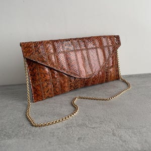 Può includere: Una pochette marrone con motivo a pelle di serpente e una tracolla a catena dorata. La borsa rettangolare ha una chiusura a patta ed è realizzata in materiale cucito e strutturato. La catena è in metallo dorato delicato.