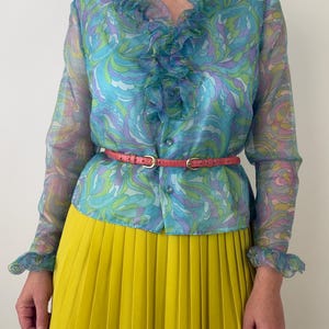May include: A long-sleeved, sheer blouse featuring a vibrant blue, purple, and green swirling pattern with a ruffled collar and cuffs. The blouse is buttoned down the front and cinched at the waist with a thin, textured coral-coloured belt. A bright yellow pleated skirt is worn below the blouse.