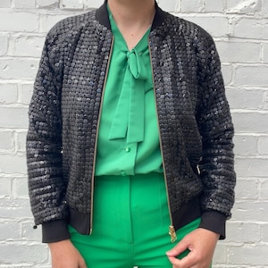 May include: A black sequined bomber jacket with a black collar and cuffs. The jacket has a gold zipper and is worn over a green blouse and green pants. The outfit is set against a white brick wall.
