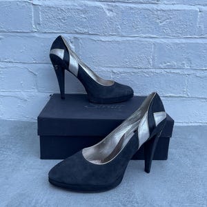 May include: A pair of black suede high-heeled shoes with silver accents. The heels are approximately 4 inches tall. The shoes are displayed on top of a black box.