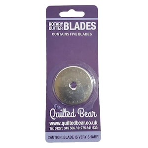 May include: A pack of five rotary cutter blades for quilting. The pack is purple and features the text "Rotary Cutter Blades" and "Contains Five Blades". The pack also includes the text "The Quilted Bear" and a website address. A warning label reads "Caution: Blade is very sharp!"
