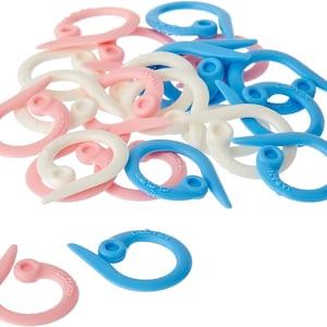 May include: A pile of plastic stitch markers in pink, white and blue. The markers are shaped like a loop with a pointed end. The markers are labelled "Clover" and "Japan".