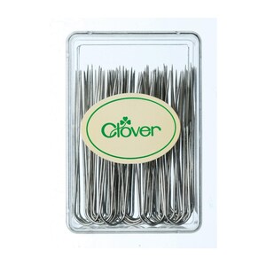 May include: A clear plastic container filled with silver-coloured Clover brand curved needles. The container has a green and cream-coloured oval label with the Clover logo and brand name.