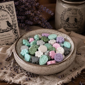 May include: A speckled ceramic bowl filled with small, molded clay shapes in shades of green, pink, white, and gray. Some pieces are shaped like flowers and leaves, while others are round with the text "be kindly" or "the kindly" imprinted.