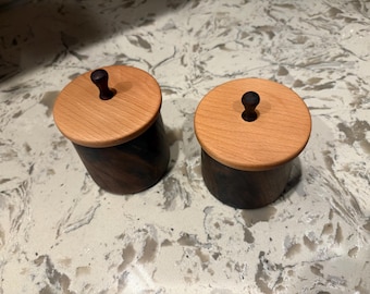 Op de afbeelding: Twee kleine, handgemaakte houten containers met deksels. De deksels zijn lichtgekleurd hout en de lichamen zijn donkerder hout. Elk heeft een kleine, donkere knop bovenop. De containers zijn waarschijnlijk voor kruiden of kleine items.