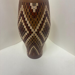 May include: A handcrafted wooden vase with a unique woven design. The vase features a combination of light and dark brown wood pieces, creating a geometric pattern. The top and bottom are a solid, darker brown. A decorative piece for the home.