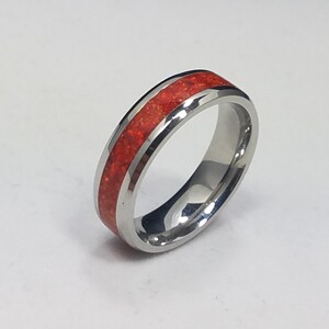May include: A polished silver-toned metal ring featuring a central inlay of vibrant red crushed material. The band has a smooth, reflective finish with beveled edges, showcasing a continuous, textured red inlay that wraps around the circumference.