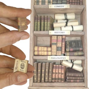 May include: Miniature wooden bookshelf filled with tiny books and scrolls. The shelves are labeled "COMEDY", "HISTORY", and "PHILOSOPHY". A small book is held in a person's hand, open to a page with text and an illustration.
