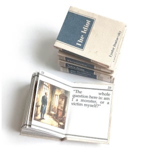 May include: A miniature book open to page 39, showing an illustration of a man in a room. The text reads "The whole question here is: am I a monster, or a victim myself?" The book cover is visible on the top of the stack, and reads "The Idiot" by Fyodor Dostoevsky.