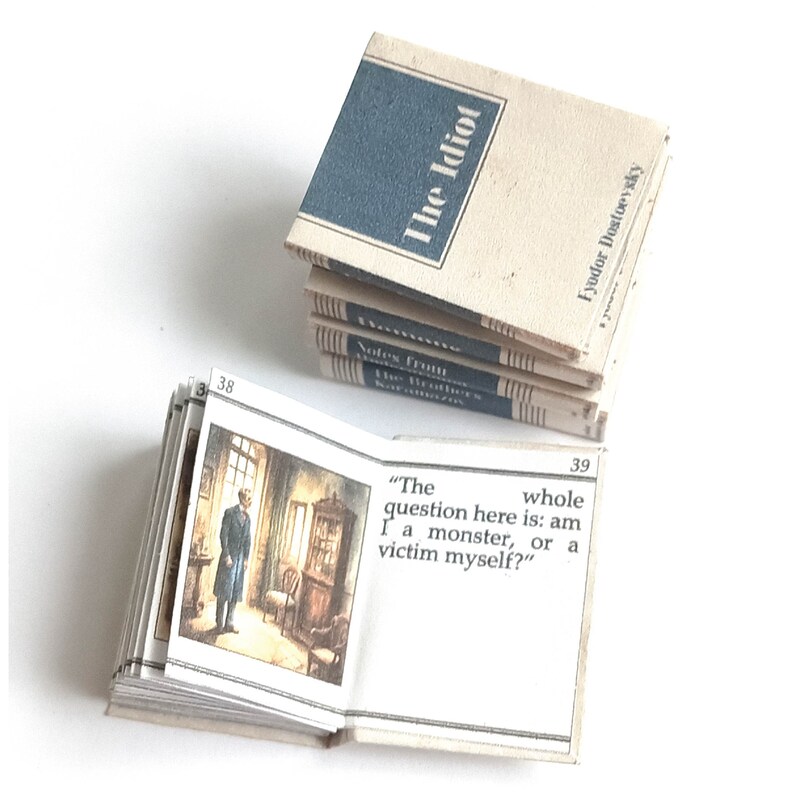 May include: A miniature book open to page 39, showing an illustration of a man in a room. The text reads "The whole question here is: am I a monster, or a victim myself?" The book cover is visible on the top of the stack, and reads "The Idiot" by Fyodor Dostoevsky.
