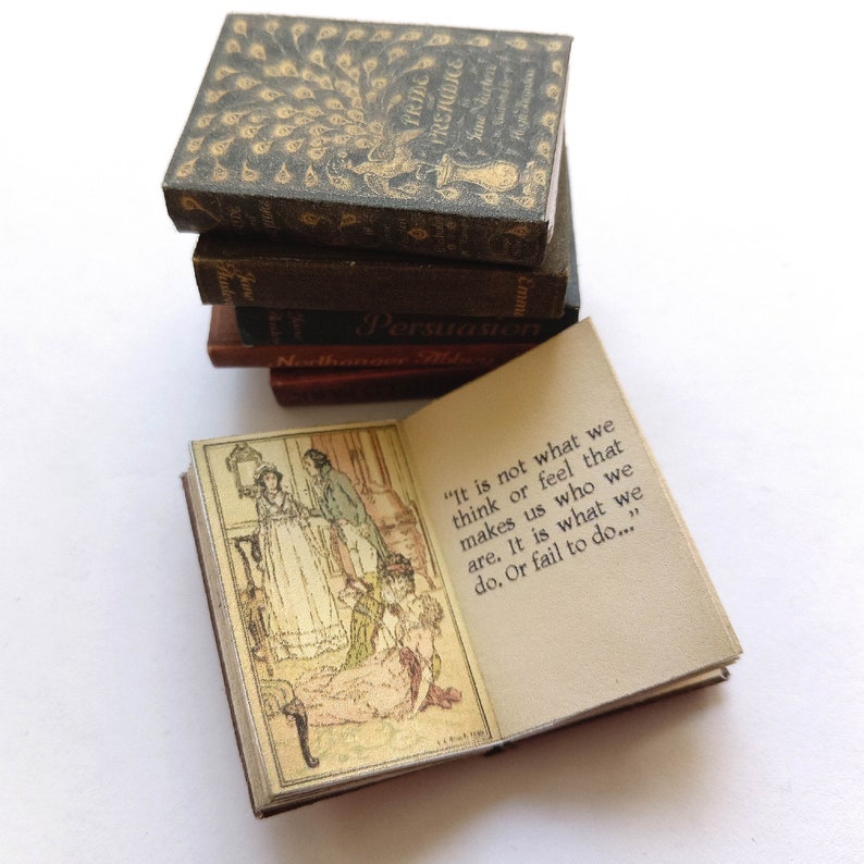 Op de afbeelding: Een stapel miniatuurboeken met een citaat uit Jane Austens *Overtuiging* op het open boek. Het citaat luidt: "Het is niet wat we denken of voelen dat ons maakt wie we zijn. Het is wat we doen. Of nalaten te doen..."