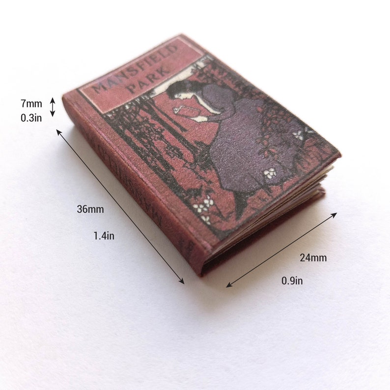 Op de afbeelding: Een miniatuurboek getiteld "Mansfield Park" met een rode kaft en een zwart-witte illustratie van een vrouw die in een bos leest. Het boek is 36 mm hoog en 24 mm breed.