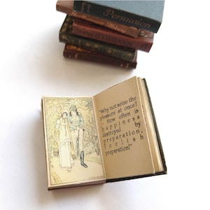 Op de afbeelding: Een klein, open boek met een groene kaft. Het boek is open op een pagina met een illustratie van twee mensen die in een bos wandelen. De tekst op de pagina luidt: "Waarom niet meteen van het plezier genieten? Hoe vaak wordt geluk vernietigd door voorbereiding, door dwaze voorbereiding!"