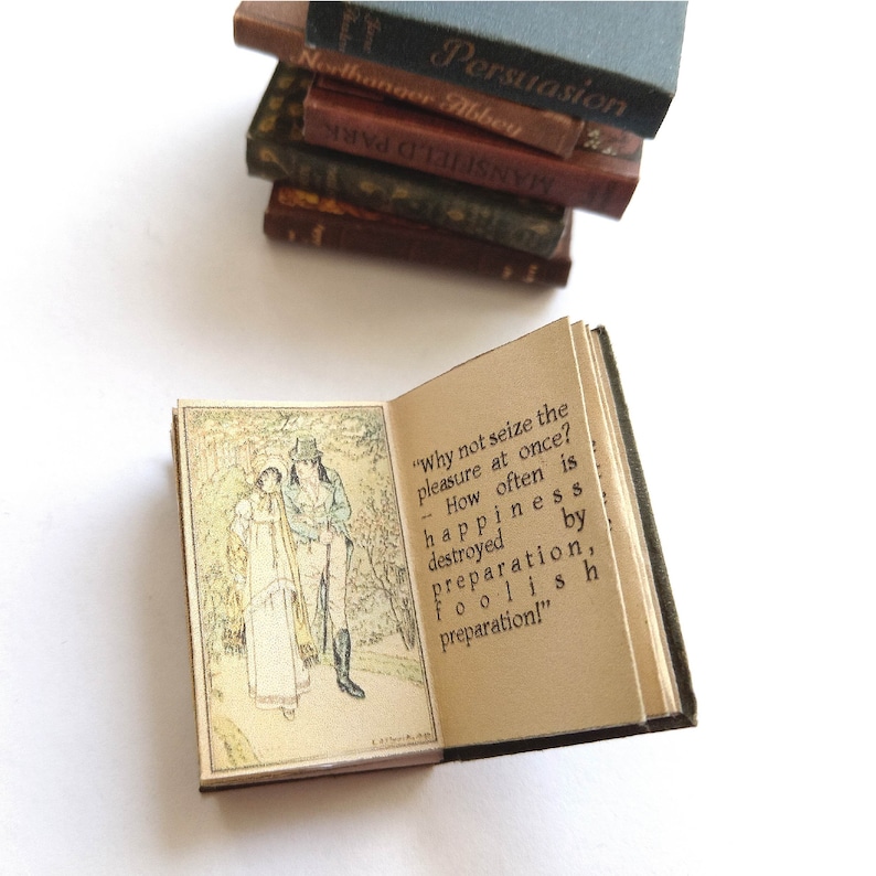 Op de afbeelding: Een klein, open boek met een groene kaft. Het boek is open op een pagina met een illustratie van twee mensen die in een bos wandelen. De tekst op de pagina luidt: "Waarom niet meteen van het plezier genieten? Hoe vaak wordt geluk vernietigd door voorbereiding, door dwaze voorbereiding!"