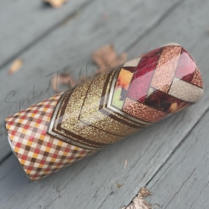 May include: A cylindrical tumbler with a decorative design. The tumbler features a plaid pattern in shades of brown, red, and yellow, transitioning to a gold glitter chevron pattern. The top section has a geometric pattern with red and gold glitter accents. The text "Spiked Tumblers" is visible.