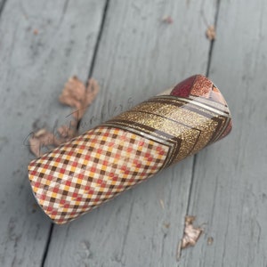 May include: A fall-themed tumbler with a plaid pattern in shades of red, brown, and yellow. The tumbler features a gold glitter band and a geometric design. The tumbler is cylindrical and appears to be made of stainless steel.