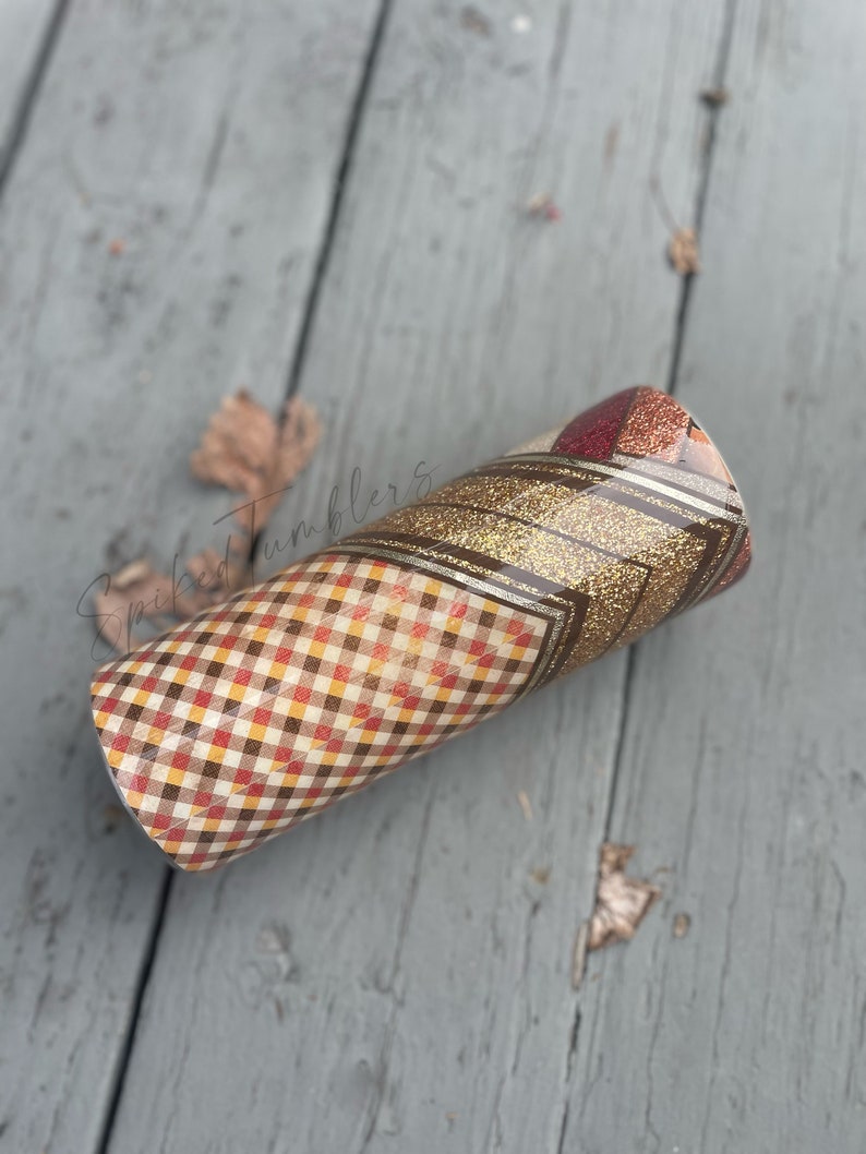 May include: A fall-themed tumbler with a plaid pattern in shades of red, brown, and yellow. The tumbler features a gold glitter band and a geometric design. The tumbler is cylindrical and appears to be made of stainless steel.