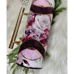 May include: A white tumbler with a gold handle and a floral design featuring purple, pink, and white flowers. The tumbler has a purple and gold glitter design.
