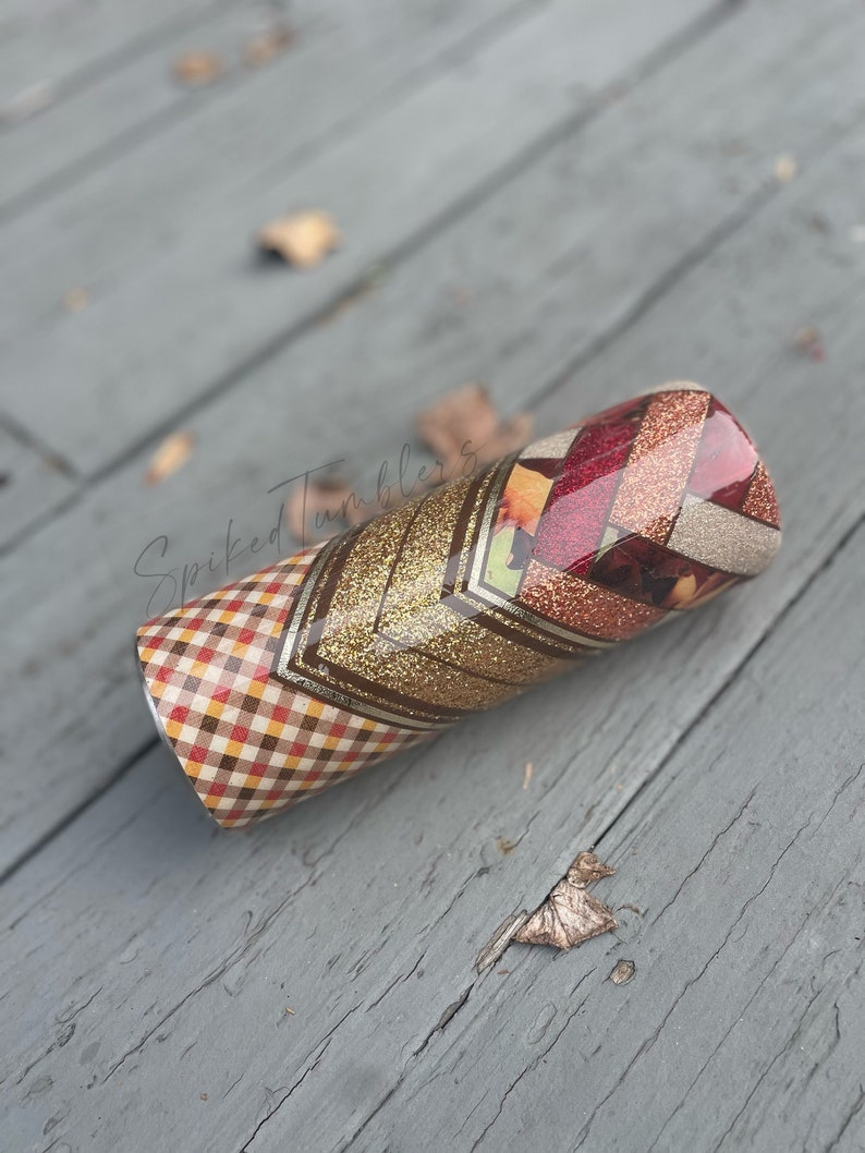 May include: A cylindrical tumbler with a decorative design. The tumbler features a plaid pattern in shades of brown, red, and yellow, transitioning to a gold glitter chevron pattern. The top section has a geometric pattern with red and gold glitter accents. The text "Spiked Tumblers" is visible.