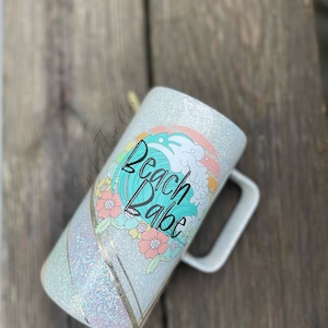 May include: White glitter tumbler with a turquoise wave design and the text "Beach Babe". The tumbler has a white handle and a gold stripe.