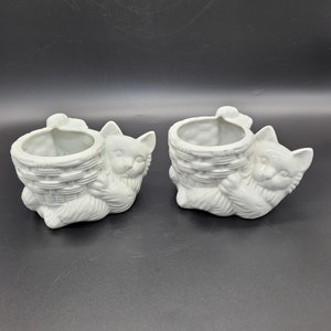 May include: Two white ceramic cat planter pots. Each pot is shaped like a cat holding a basket in its paws. The cats are facing forward and have a sweet expression on their faces.