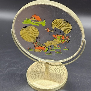 May include: A gold-colored tabletop vanity mirror with a round, mirrored surface. The mirror features a cartoon image of two children on a seesaw in a green field with a small house in the background. The mirror has a decorative base with a gold paisley pattern.