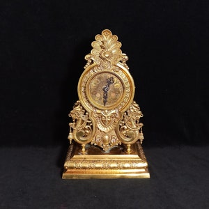May include: An ornate gold-toned clock with a Roman numeral face and a decorative base. The clock features a detailed design with a central face surrounded by intricate carvings.