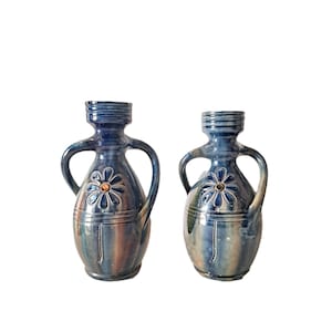 Könnte beinhalten: Zwei blaue Keramikvasen mit Henkeln. Jede Vase hat ein Blumendesign in der Mitte und eine strukturierte Oberfläche. Die Vasen haben ein rustikales, handwerkliches Aussehen mit einer Mischung aus Blau-, Braun- und Grüntönen.