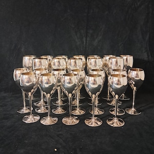 Könnte beinhalten: Eine Sammlung silberfarbener Weingläser. Die Gläser haben eine runde Schale, einen schlanken Stiel und einen kreisförmigen Fuß. Die Gläser sind in Reihen angeordnet und reflektieren das Licht. Der Hintergrund ist dunkel und einfarbig.