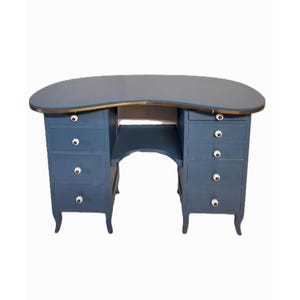 Op de afbeelding: Een blauwe bureau met een gebogen bovenkant en messing afwerking. De bureau heeft meerdere lades met witte knoppen en een kleine plank. De bureau is in vintage stijl en zou geschikt zijn voor een slaapkamer of kantoor. Meet ongeveer 150 cm lang.