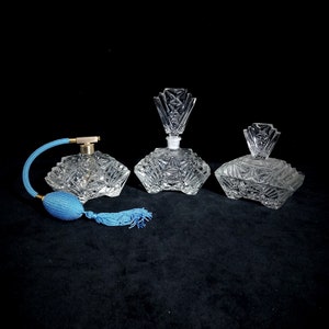 Puede incluir: Tres frascos de perfume de vidrio transparente con intrincados diseños de vidrio cortado. El frasco de la izquierda tiene un atomizador azul y una borla. Los frascos están dispuestos sobre una superficie negra.