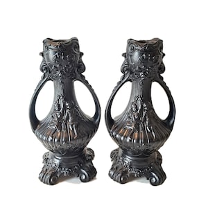May include: A pair of black decorative vases with intricate designs. Each vase has a broad base, a central section with sculpted figures, and a flared top with handles. The vases are symmetrical and set against a white backdrop.