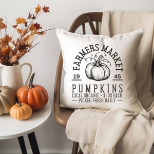 Könnte beinhalten: Ein weißes quadratisches Kissen mit einem schwarzen Aufdruck "FARMERS MARKET PUMPKINS" und einer Kürbisillustration. Das Kissen liegt auf einem Holzstuhl mit einer beigen Decke. Kürbisse und Herbstlaub liegen auf einem weißen Tisch.