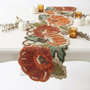 May include: A long, narrow table runner featuring intricate beadwork in shades of orange, brown, and green. The design displays multiple pumpkin motifs surrounded by detailed leaf patterns, all crafted with small, reflective glass beads.