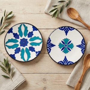 May include: Two round ceramic plates featuring a white base with hand-painted blue and teal floral patterns. The plates have a dark rim and are arranged alongside two wooden spoons and green olive branches on a light wooden surface.