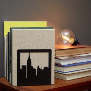 May include: Black metal bookends shaped like a city skyline, holding up a stack of books. The bookends are on a wooden table with a light bulb in the background.