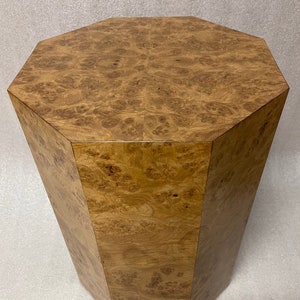 May include: A hexagonal pedestal stand featuring a light brown wood burl veneer with a complex, swirling grain pattern. The structure has flat, vertical sides and a flat top surface, showcasing a polished finish that highlights the natural wood texture.