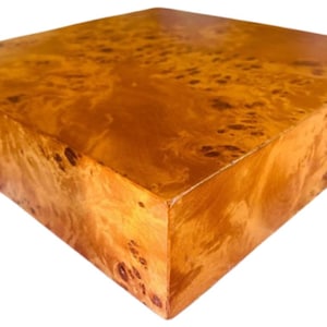 May include: A square wooden box with a warm, golden-brown finish. The wood grain features a unique, swirling pattern with darker brown accents. The box has a smooth, polished surface and a simple, geometric design.