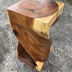 May include: A handcrafted wooden side table with a unique, twisted design. The table is made from a light and dark brown wood, with visible wood grain and a smooth, polished finish. The top is square, and the base is also square. The table is approximately 18 inches tall.