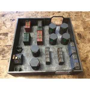 Könnte beinhalten: Ein Miniaturmodell eines futuristischen Raumes mit einem grauen Fliesenboden, schwarzen und grauen Wänden und verschiedenen schwarzen, grauen, grünen und roten Möbelstücken. Der Raum hat ein großes Fenster mit einem schwarzen Rahmen und eine Tür mit einem schwarzen Rahmen. Der Raum wird von einer einzigen Leuchte an der Decke beleuchtet.