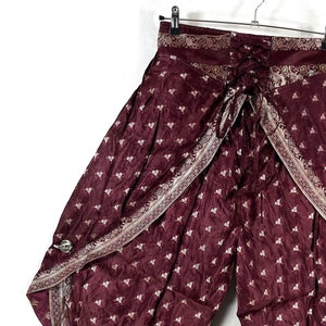 May include: Burgundy palazzo pants with a floral pattern and decorative trim. The wide-leg pants have a wrap-around design with a tie closure at the waist. The fabric has a slight sheen, and the trim is a lighter shade of burgundy.