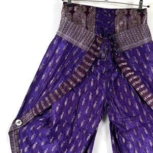 May include: Purple wide-leg pants with a paisley pattern and silver accents. The high-waisted pants feature a decorative waistband and side button closures. The fabric has a shimmering effect, and the pants are hanging on a metal hanger.