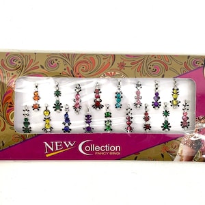 May include: A sheet of colourful, jewel-toned, adhesive bindis with silver settings. The bindis are arranged in rows on a pink and gold background with the text "NEW Collection FANCY BINDI".