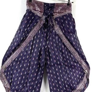 May include: Wide-leg, dark purple pants with a paisley pattern and decorative trim. The pants have a lace-up back and are hanging from a metal hanger. The fabric has a slight sheen.